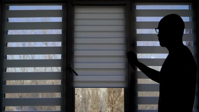 the silhouette of an adult man who opens the blinds in his apartment, the pearson wants to see the weather on the street