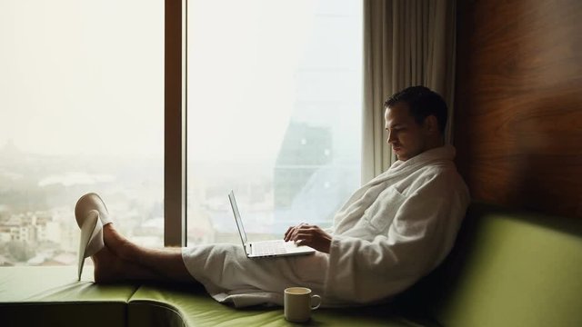 Young smiling businessman working on laptop computer wearing white bath robe sitting near window with cup of coffee looking at sunrise city view. Motivation concept