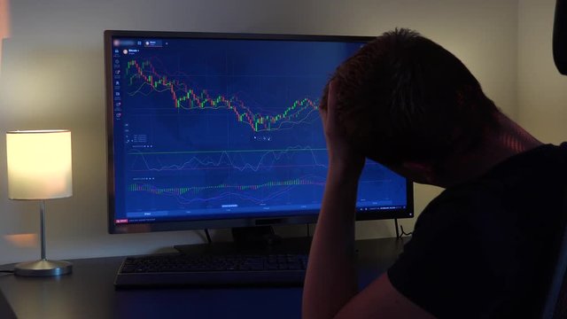A man sits at a table, looks at a candle chart on a computer screen and gets frustrated