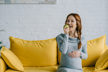 happy attractive pregnant woman eating apple in living room