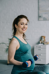 Fototapeta na wymiar smiling pregnant woman exercising with dumbbells and looking at camera