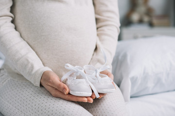 cropped image of pregnant woman holding newborn shoes in bedroom