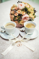gold wedding rings lie on the table next to two cups of coffee and wedding bouquet