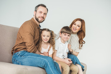 cheerful family with two children sitting on couch and smiling at camera on grey