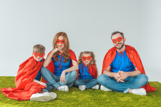 super family in masks and cloaks sitting on green grass and smiling at camera on grey
