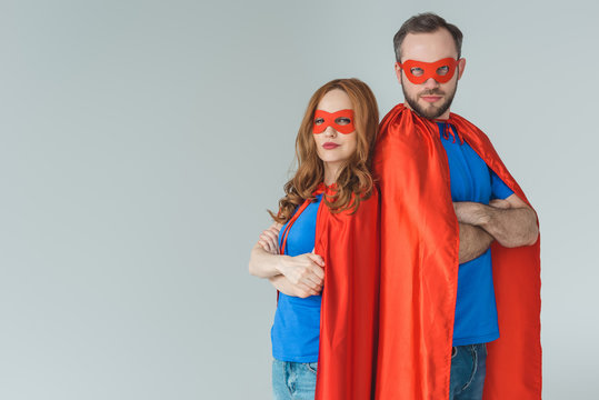 super couple in masks and cloaks standing with crossed arms and looking at camera isolated on grey