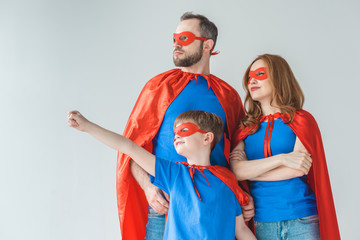 family of superheroes in masks and cloaks standing together and looking away isolated on grey