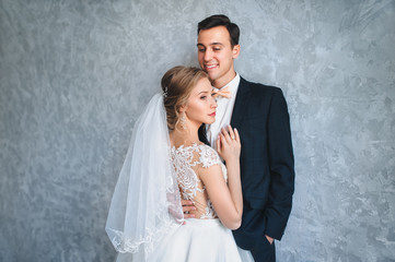 A young bridegroom embraces a beautiful bride in a lace dress near a gray wall. Portrait of a...