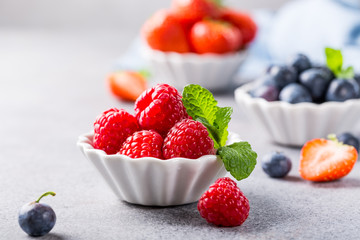 Fresh ripe raspberries, strawberries and bluebetties in white bowls on light gray concrete background. Healthy food concept with copy space.