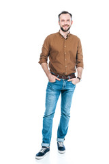 Fototapeta premium full length view of handsome bearded man standing with hands in pockets and smiling at camera isolated on white