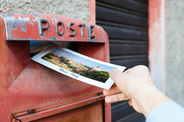 Hand drops a postcard in a red mailbox in the Rome, Italy. The postcard shows a panorama of Rome.