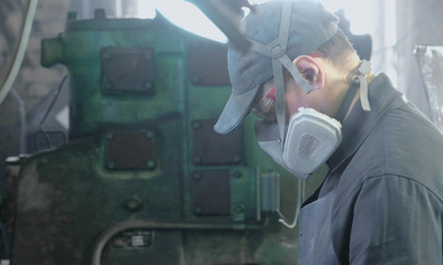 Worker performs his job in a protective mask on his face in the shop among the equipment. - obrazy, fototapety, plakaty
