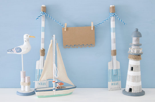 nautical concept with wooden decorative boat oars and empty hanging note on a string next to lighthouse, seagull and boat over blue background.