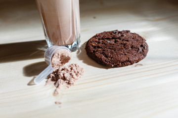 Mixed milk chocolate with whey protein shake in glass cup, scoop with protein and chocolate cookie on wooden table. Dietary snack for bodybuilding. Protein casein, geiner, hydrolysate after training