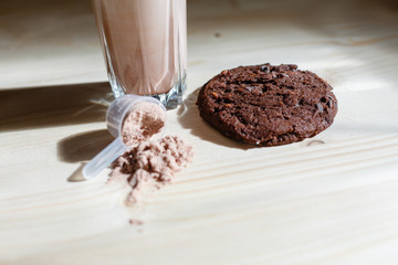 Mixed milk chocolate with whey protein shake in glass cup, scoop with protein and chocolate cookie on wooden table. Dietary snack for bodybuilding. Protein casein, geiner, hydrolysate after training
