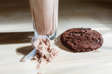 Mixed milk chocolate with whey protein shake in glass cup, scoop with protein and chocolate cookie on wooden table. Dietary snack for bodybuilding. Protein casein, geiner, hydrolysate after training