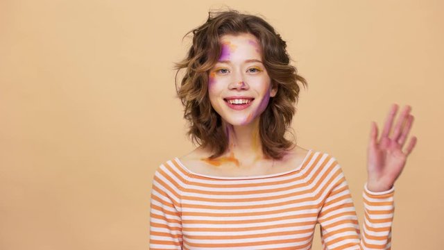 Portrait of adorable woman 20s with colorful painted spots on her face smiling and waving hand in welcoming, isolated over beige background. Body language