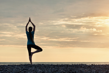 Girl at sunset practicing yoga at the seashore, back view