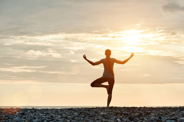 Fototapeta premium Girl at sunset practicing yoga at the seashore, back view