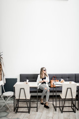 Stylish business woman in white jacket sitting during the coffee break in the modern cafe interior. Wide view with copy space on the wall