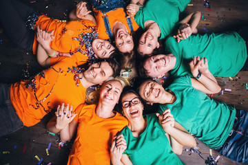 Fototapeta premium Happy friends lying together in circle looking at camera, keeping hands near mouth and smiling while lying on wooden floor.. Concepts about friendship,lifestyle,unity,business and teamwork.
