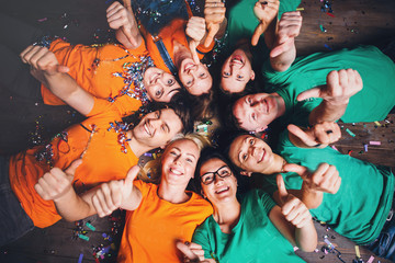 Fototapeta premium Happy friends lying together in circle looking at camera, keeping hands near mouth and smiling while lying on wooden floor.. Concepts about friendship,lifestyle,unity,business and teamwork.