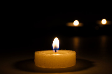 Fototapeta na wymiar The flame of a burning round candle casts shadows on the surface, on the back of a blurry background two lit candles.