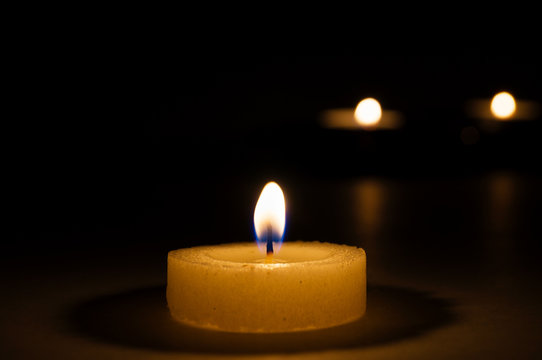 The flame of a burning round candle casts shadows on the surface, on the back of a blurry background two lit candles.