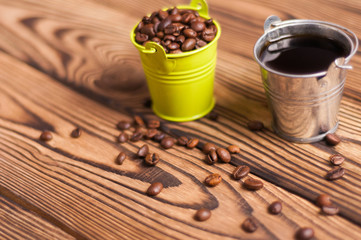 Full zinked bucket of black coffee near scattered lot of roasted beans and green metal bucket with handle full of coffee beans on old worn wooden table