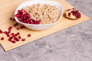 Cooked yummy warm oatmeal and heap of ripe fresh pomegranate seeds in white ceramic bowl near piece of pomegranate on clean bamboo rug on worn gray scratched concrete with copy space