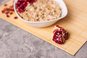 Cooked yummy warm oatmeal and heap of ripe fresh pomegranate seeds in white ceramic bowl near piece of pomegranate on clean bamboo rug on worn gray scratched concrete with copy space