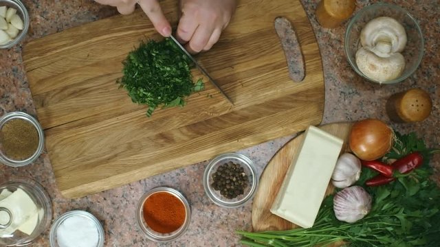 Fast motion top view of unrecognizable cook with knife cutting fresh parsley on wooden board; bowls with spices and assorted ingredients placed on table