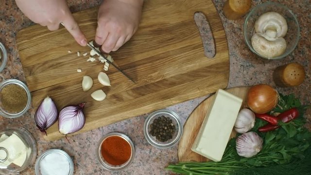 Top view of unrecognizable cook with knife chopping garlic cloves on wooden board; bowls with spices and assorted ingredients placed on table