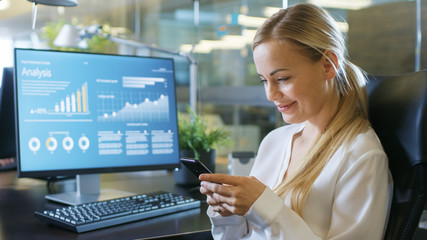 In the Office Attractive Woman Swirles in Chair, Smiles and Uses Mobile Phone. Her Desktop Computer Screen Shows Infographics with Statistical Growth of Her Company's Shares. Stylish Room Setting.