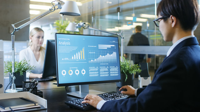 In The Office Businessman Works At His Desk, He's Using Personal Computer With Statistical Information Showing On It's Screen. His Colleague Sets Opposite To Him.