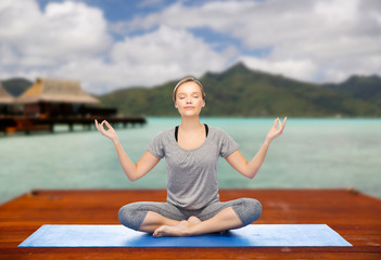 fitness, meditation and healthy lifestyle concept - happy woman making yoga lotus pose on wooden pier over island beach and bungalow background