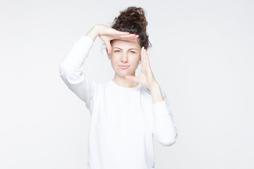 Indoor studio shot of  young attractive European female with curly hair bun smiling at camera and gesturing finger frame, while standing against isolated white wall background. Body language concept.