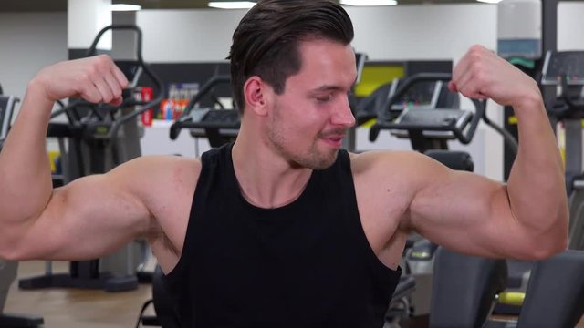 A young fit man smiles and shows off his biceps for the camera in a gym - closeup