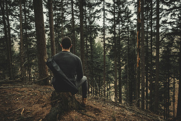Fototapeta premium Young man (camper and hiker) sitting on the stump on peak in coniferous forest and enjoying amazing view. Man - Alone in forest. Resting and relaxing in forest.