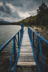 Fototapeta premium Mole (pier) on the scandinavian lake with motorboat on side and misty hills. Blue wooden bridge in forest in autumn time with blue lake. Lake for fishing with pier and powerboat. Foggy lake with hills