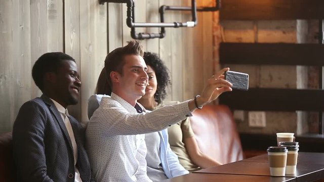 Diverse happy friends making selfie on smartphone together in cafe, caucasian guy holding phone taking picture or shooting video of hanging out with multi-ethnic african young people in coffeehouse