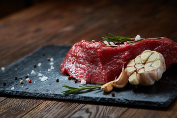 Raw meat beef steaks on black slate board with spices, garlic and rosemary over wooden background, selective focus
