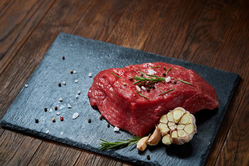 Raw meat beef steaks on black slate board with spices, garlic and rosemary over wooden background, selective focus