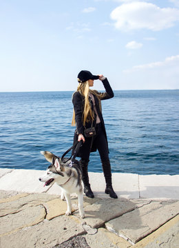 young sexy blonde sunny girl in black cap is standing fashion and posing with cute husky on the blue sea background