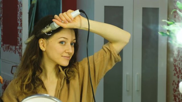 Young beautiful girl looks in the mirror and curls her hair on a curling iron in the dressing room, the girl is getting ready for a date