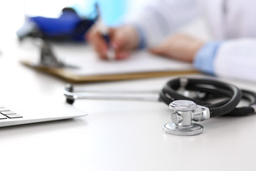 Closeup of stethoscope. Female doctor fills up medical form while sitting at the desk in hospital. Healthcare, workplace and cardiology in medicine concept