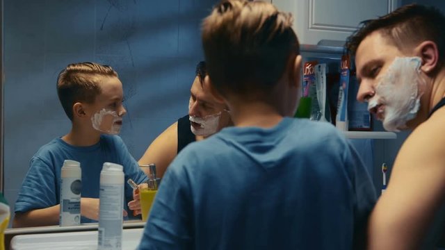 Young boy learning to shave helped by his father as they peer at their reflections in a bathroom mirror with chins covered with shaving foam as the youngster tries out a razor.
