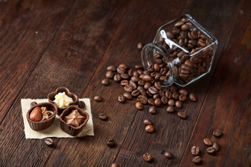 Side view of overturned glass jar with coffee beans and chocolate candies on wooden background,...