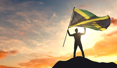 Jamaica flag being waved by a man celebrating success at the top of a mountain. 3D Rendering - obrazy, fototapety, plakaty