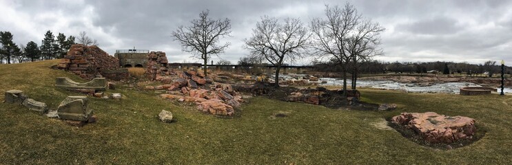 The Big Sioux River flows over rocks in Sioux Falls South Dakota with views of wildlife, ruins, park paths, train track bridge, trees and city in the surrounding area and background